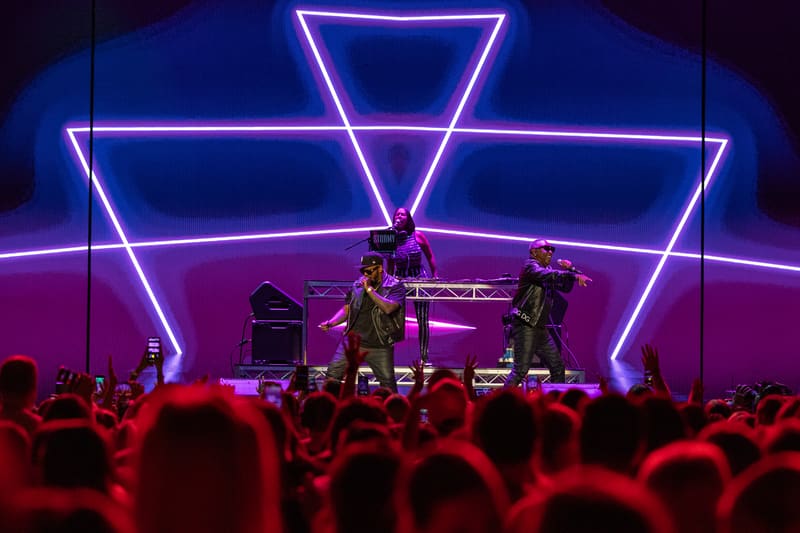 Band performing on stage framed by neon pink and blue geometric lines, audience silhouettes in foreground.