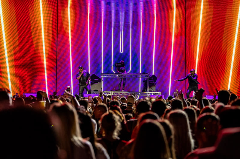 Crowd of concert-goers facing a dimly lit stage with neon purple and red vertical light installations behind silhouetted performers.