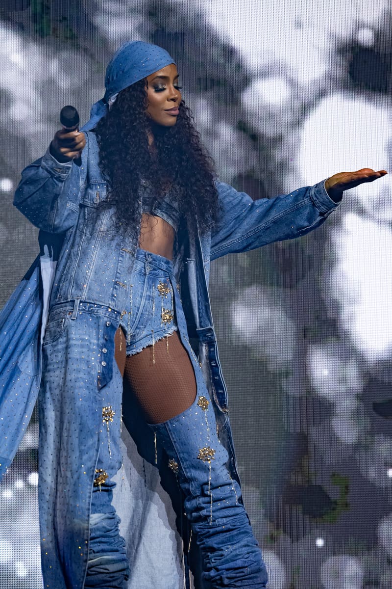 Performer in blue denim outfit holding microphone with arm extended during stage performance with bokeh lighting backdrop.