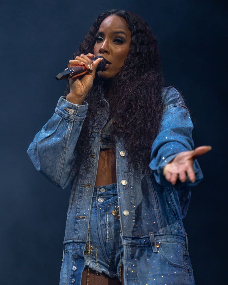 Singer performing on stage in denim jacket, holding microphone against dark blue background.
