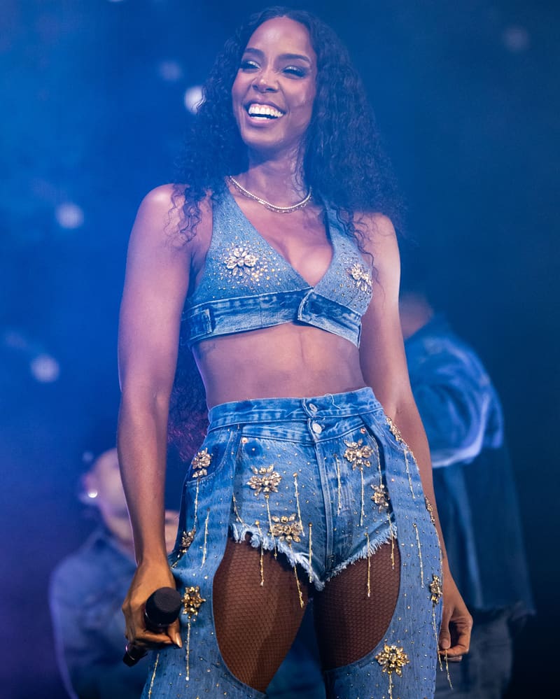 Performer on stage wearing a sequined denim crop top and distressed denim shorts, smiling under blue stage lighting.