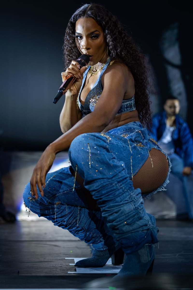 Singer performing on stage in distressed blue jeans and black top, holding microphone with long curled hair and jewellery.