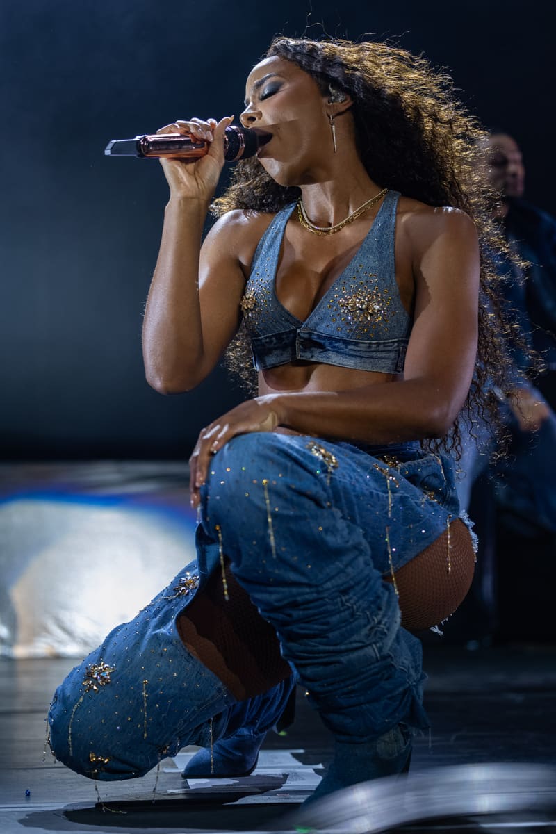 Singer performing on stage in a sparkly blue crop top and jeans, kneeling whilst holding a microphone.