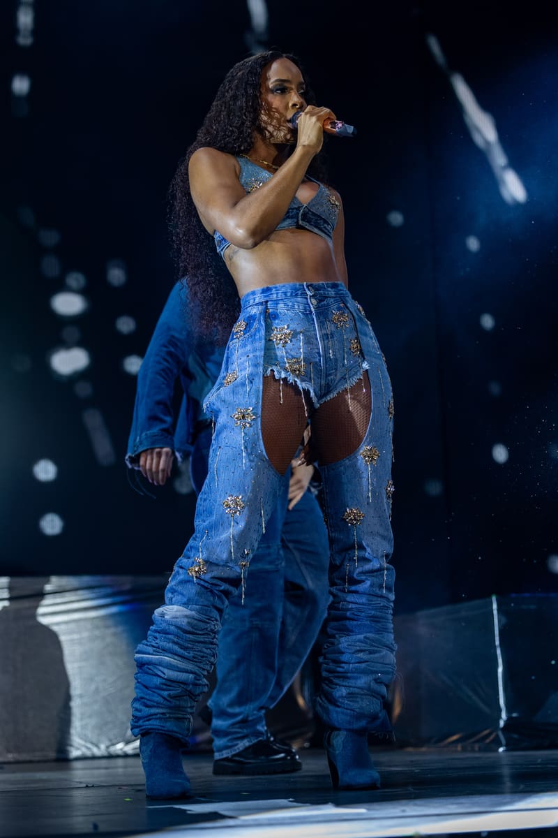 Performer singing into microphone on stage wearing blue distressed denim and crop top, with long dark hair and stage lighting behind.