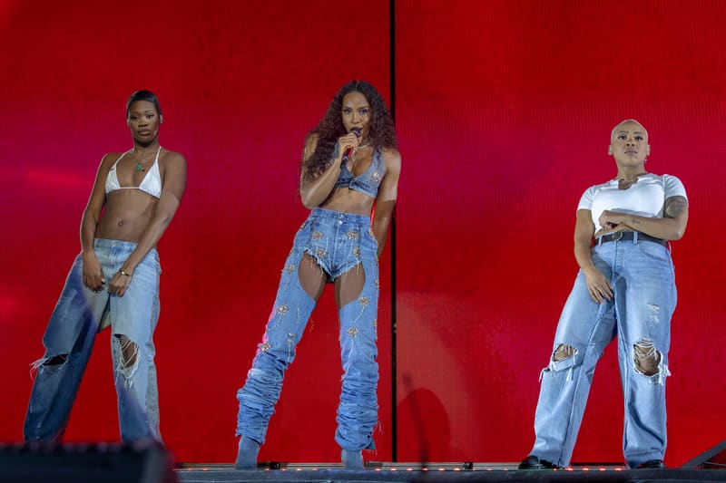 Three performers in ripped denim and coordinated crop tops stand against a vibrant red stage backdrop during a live concert performance.