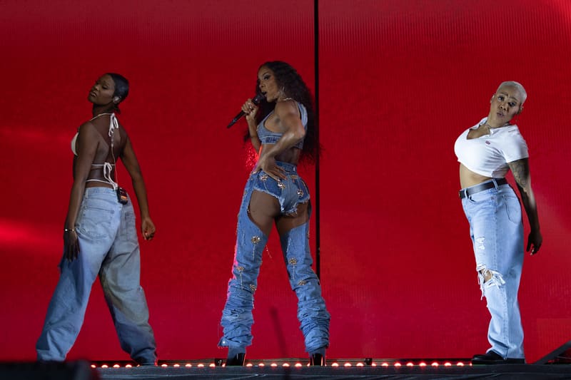 Three performers in denim and crop tops stand on a stage lit by red backdrop and footlights during a live concert performance.