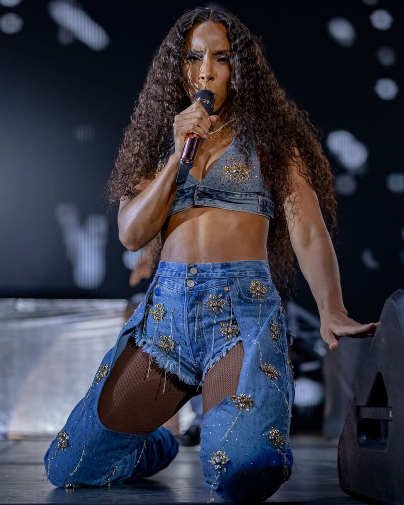 Performer with long curly hair kneels on stage wearing a denim crop top and distressed jeans, singing into a microphone.