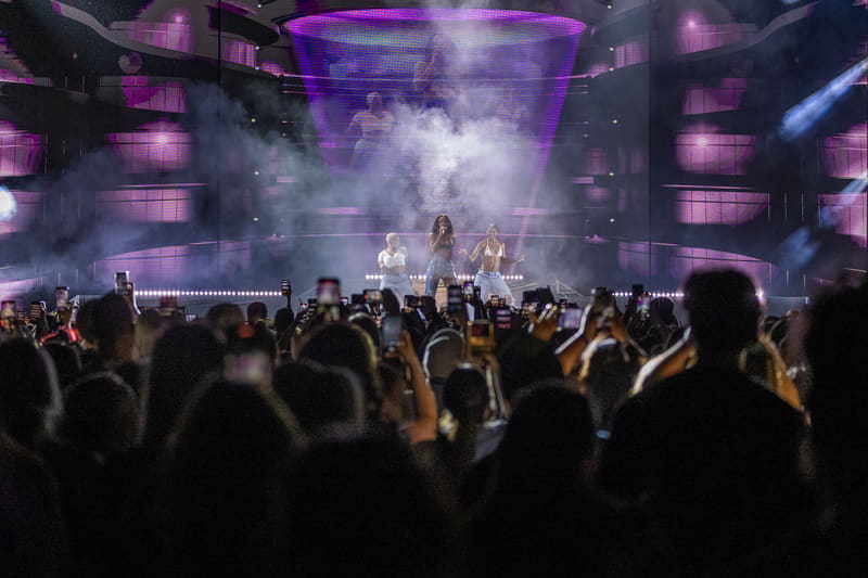 Performer on stage beneath purple lighting and smoke as a large crowd watches with phones raised during a live concert.