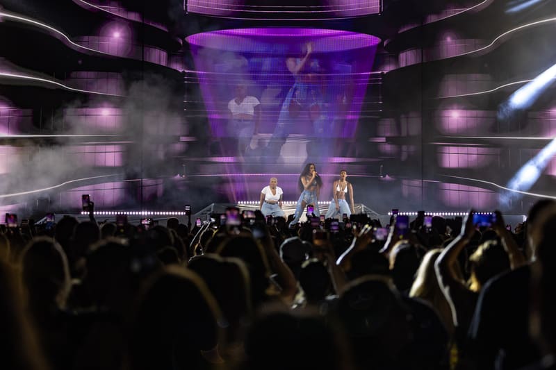 Performer on stage bathed in purple lighting amid a crowd of audience members with raised hands at a live concert.