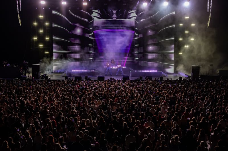 Large crowd facing an illuminated stage with purple lighting and smoke effects during a live music performance.
