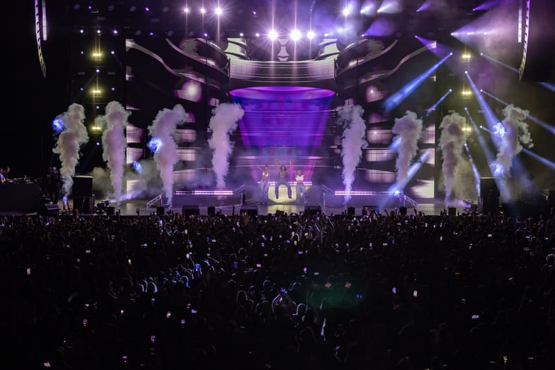 Large-scale concert stage with purple lighting, towering white columns, and crowd of spectators in foreground.
