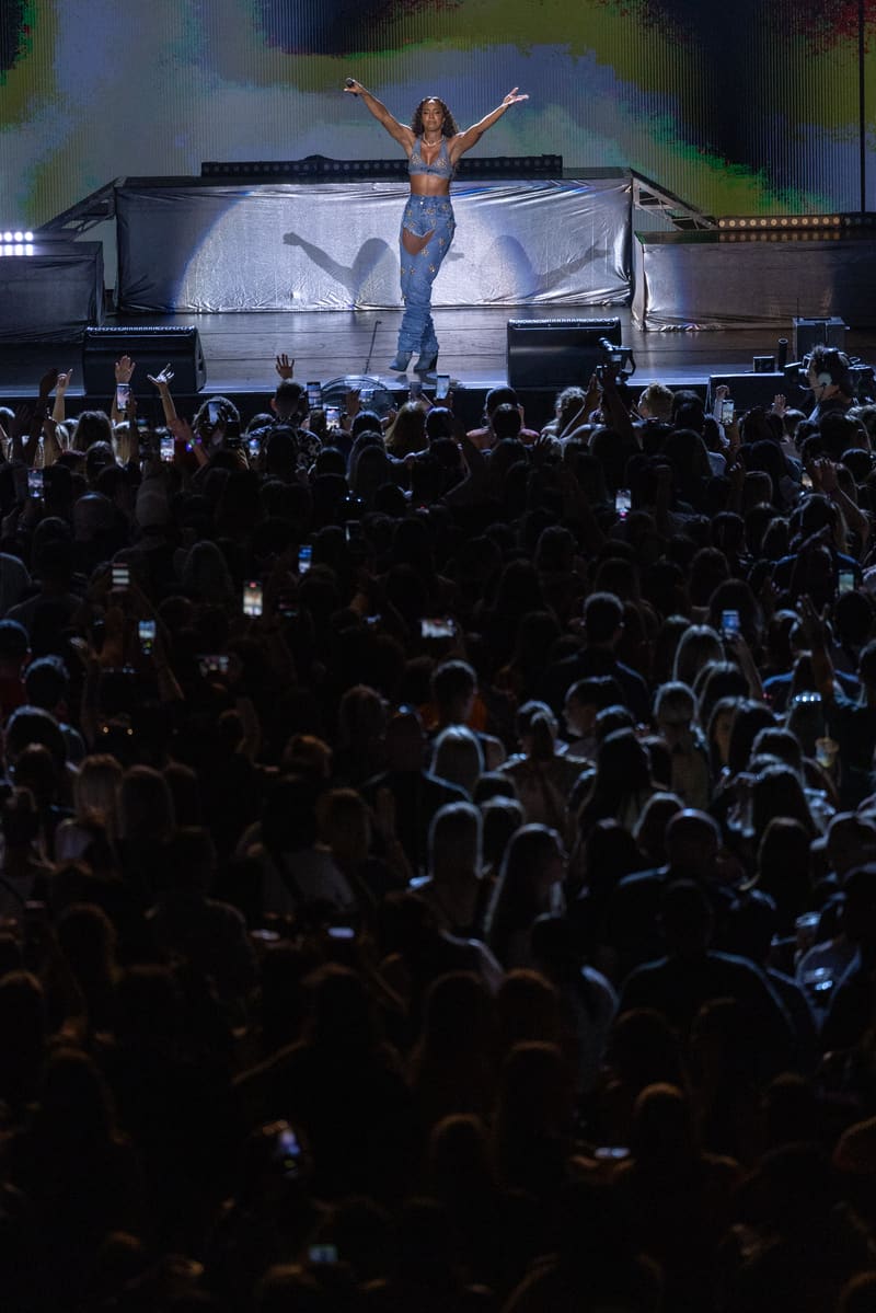 Performer in a shimmering blue outfit with arms raised on stage, surrounded by a cheering crowd holding phones.