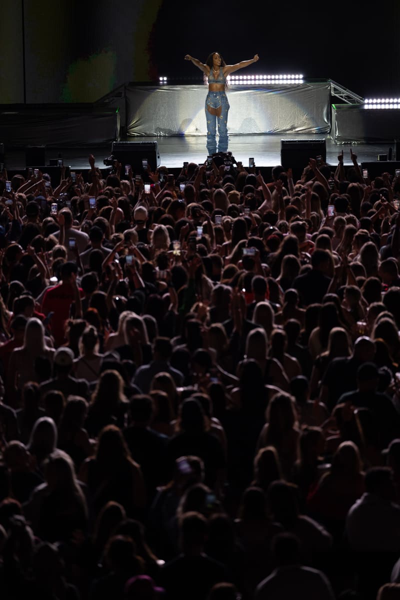 Performer on stage with arms raised above a large crowd during an indoor concert, lit by bright stage lighting.