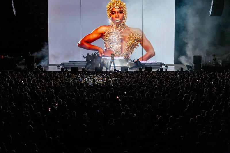 Large crowd facing a stage screen displaying a performer in a gold sequinned outfit with a striking golden afro hairstyle.