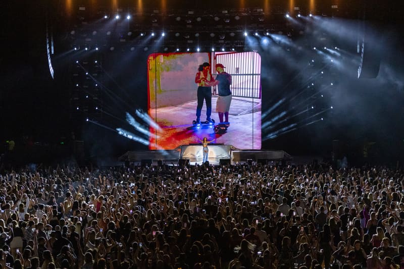 Performer on stage beneath large screen display, surrounded by thousands of audience members at concert venue.