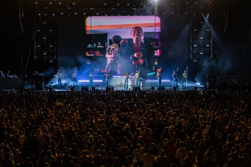 Large crowd facing a lit stage with performers and a video screen during an evening concert.