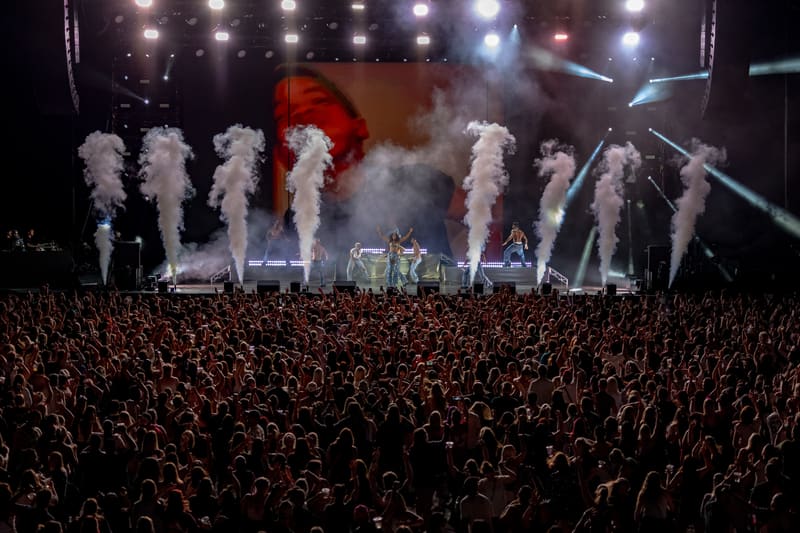 Stage performers in feathered white costumes amid pyrotechnic effects, surrounded by a dense crowd at a concert venue.