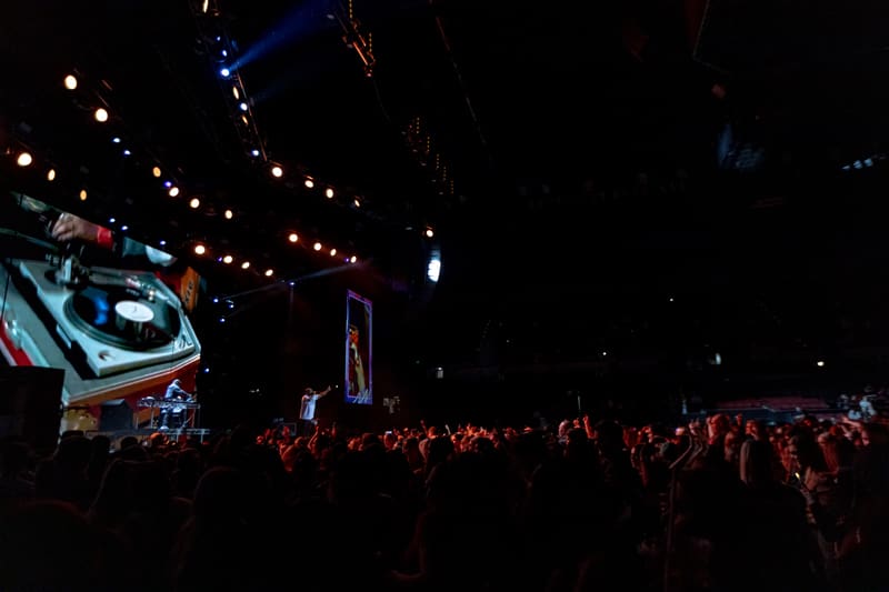 Large crowd illuminated by stage lighting at an indoor live music venue with overhead lights and projection screens.