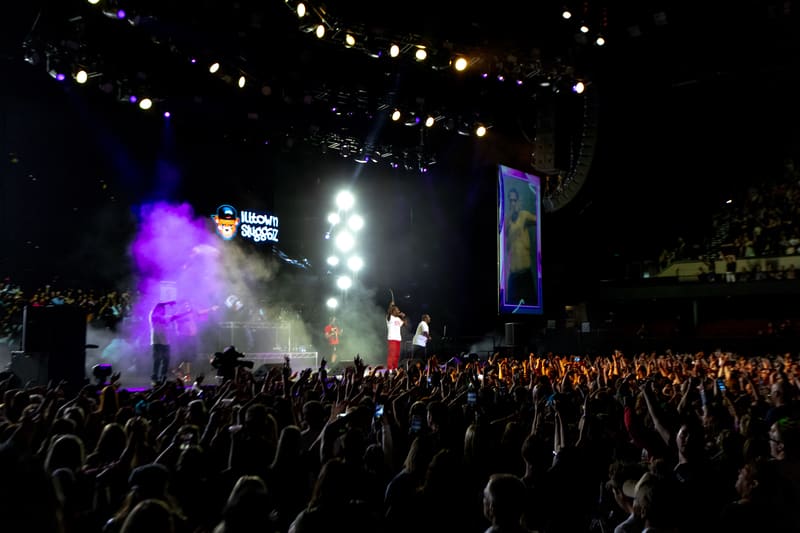 Crowd with raised hands facing a stage lit by purple and white spotlights during an outdoor music event.