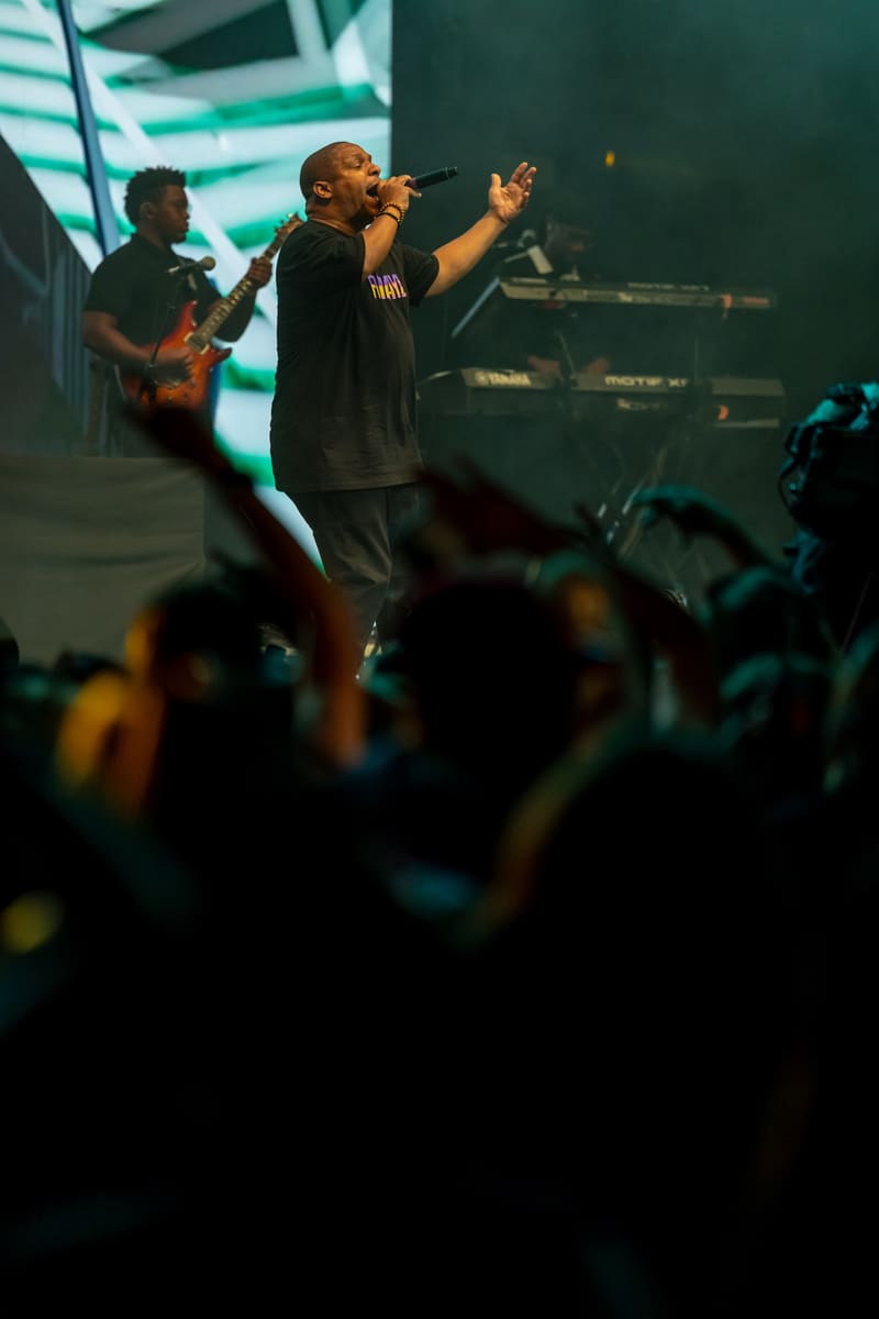 Performer rapping centre stage with arms raised, band behind, crowd silhouetted in foreground under green lighting.