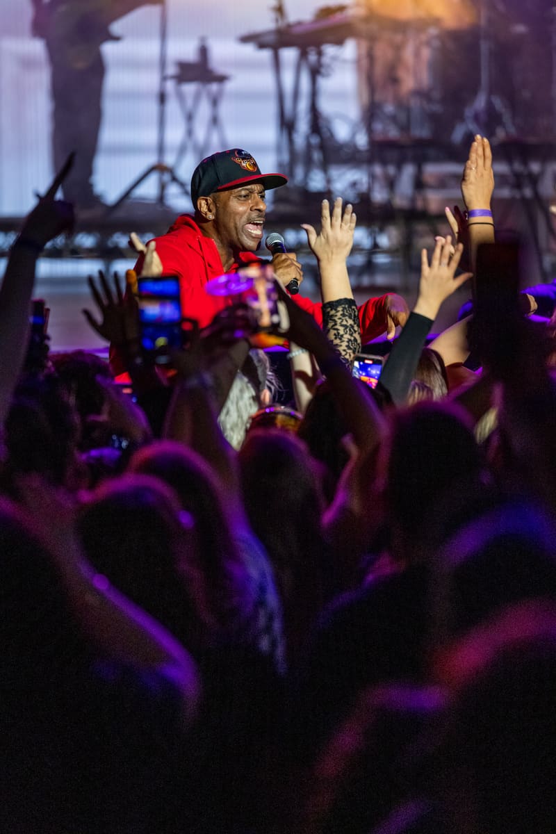 Performer in red jacket and cap holding microphone amid raised hands of engaged crowd under purple stage lighting.