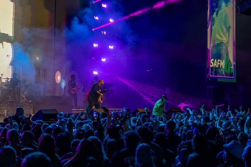 Performer on stage bathed in purple and blue lighting above a dense crowd with raised hands at a live music event.