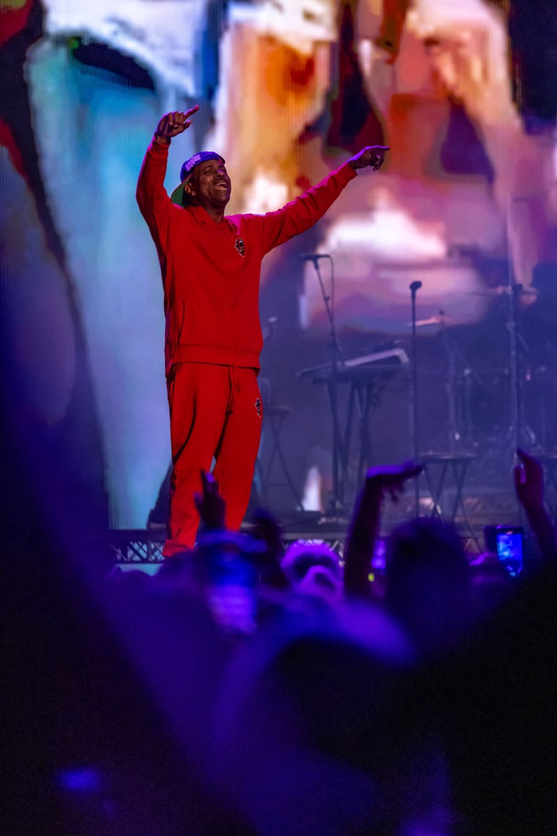 Performer in red suit with arms raised on stage amid purple lighting and crowd at live music event.