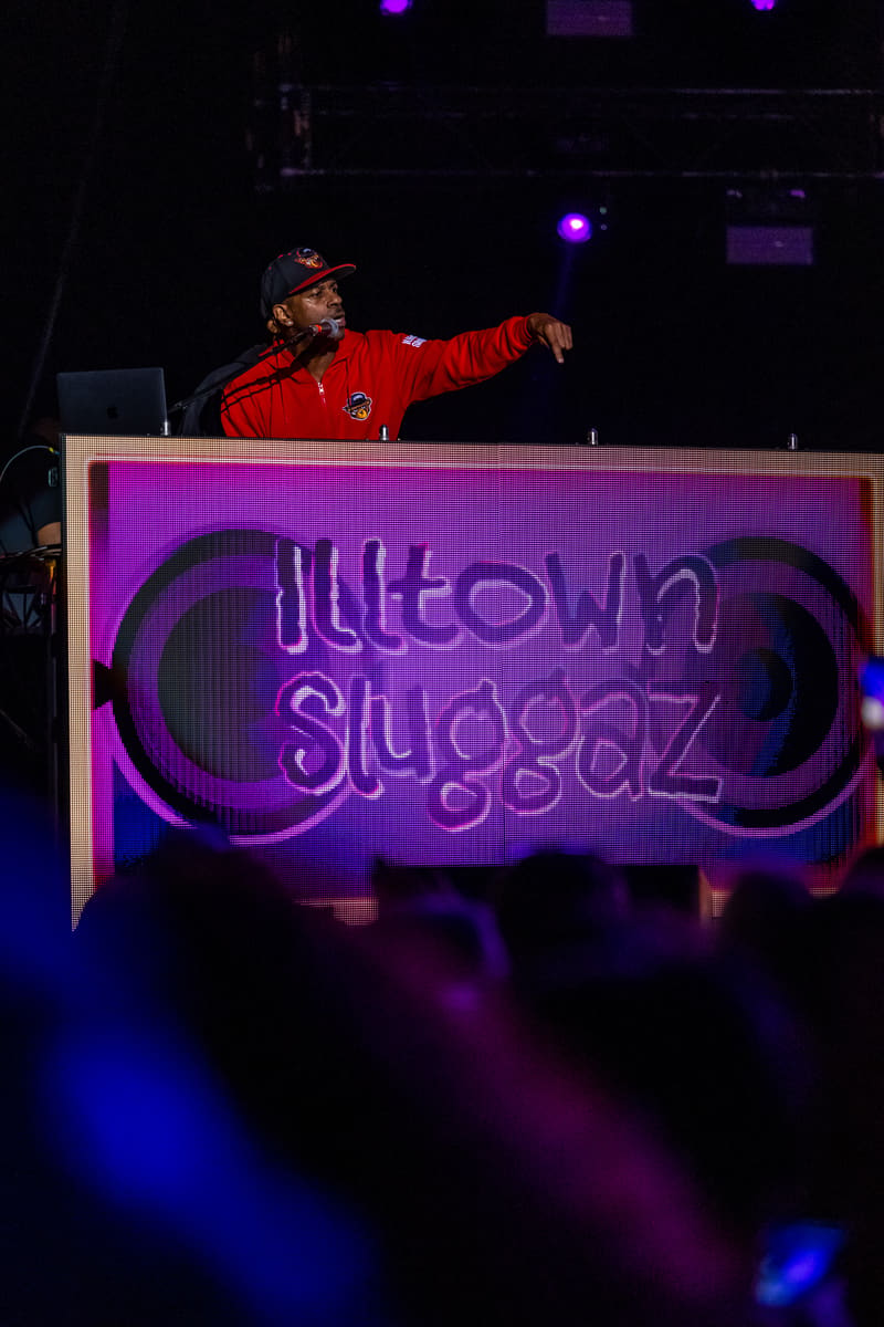 DJ in red jacket gestures over illuminated Illtown Sluggaz neon sign during live performance.