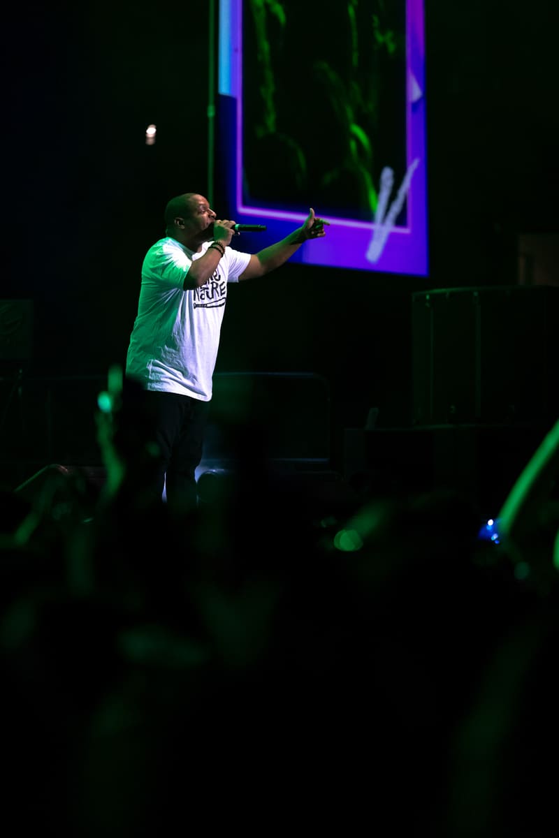 Performer in white and green shirt rapping on stage under blue and green neon lights.