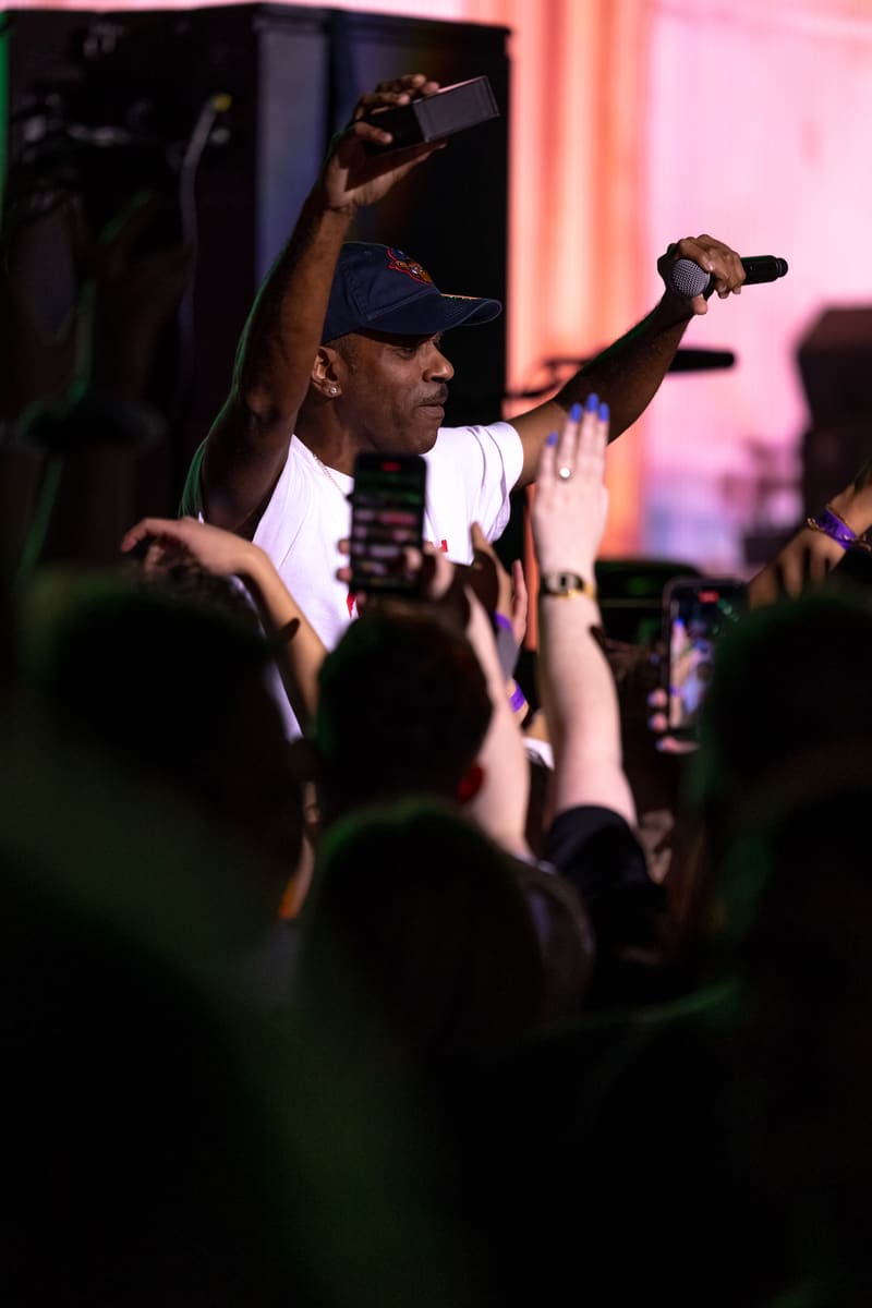 Performer rapping on stage with raised arms while audience members watch below, bathed in pink stage lighting.