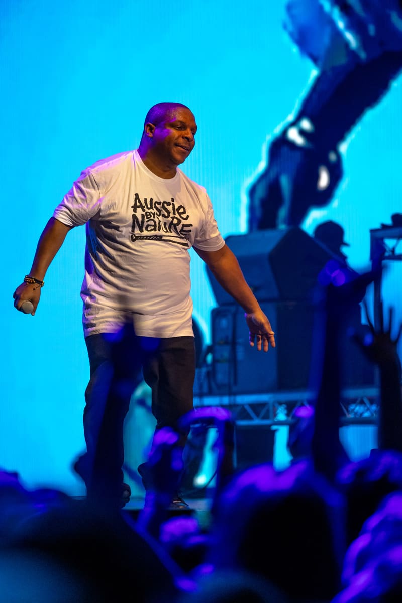 Performer on stage wearing white "Aussie Nativez" t-shirt, bathed in blue and purple stage lighting during live show.