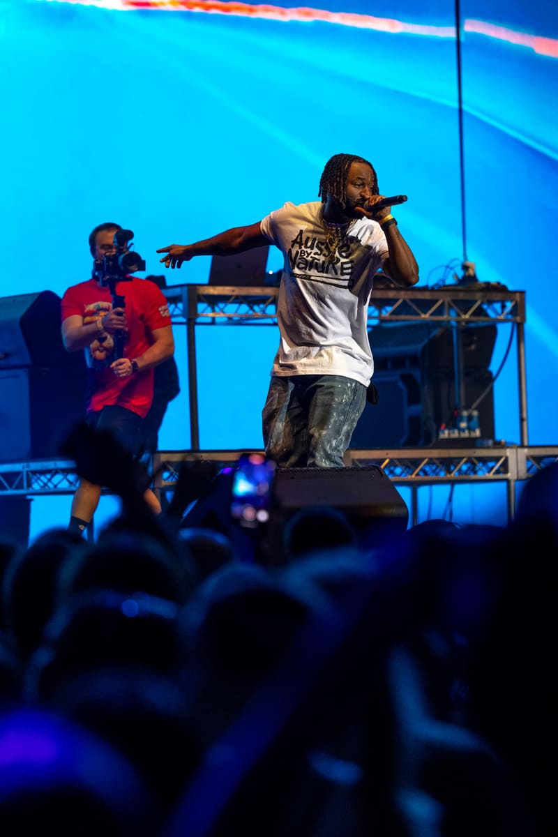 Performer in white T-shirt mid-rap on stage with blue lighting and crowd visible below.