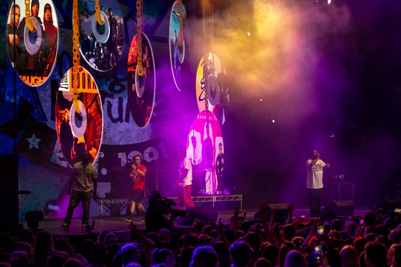 Performer illuminated by purple and pink stage lights on a concert stage with large illuminated logos overhead and silhouetted audience in foreground.