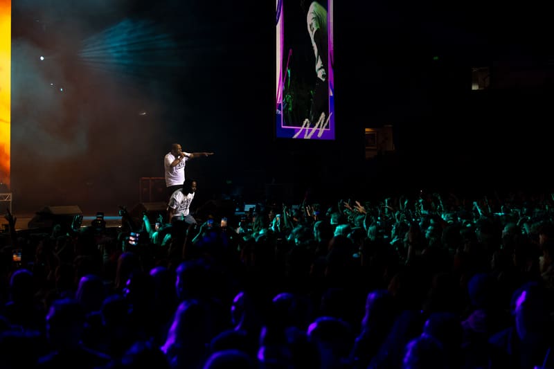 Performer on stage beneath neon signage, crowd with raised arms illuminated by blue stage lighting.