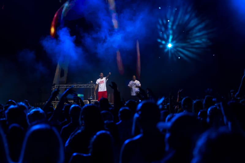 Two performers on stage in front of a crowd illuminated by blue stage lighting and pyrotechnics.