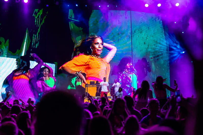 Performer in yellow shirt on stage with raised arm, surrounded by purple and blue stage lighting and silhouetted crowd.