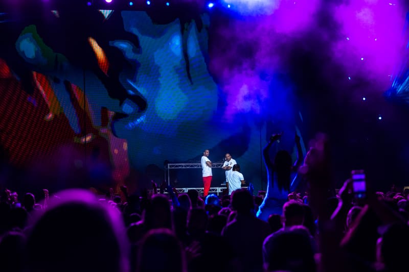 Two performers on stage beneath vibrant blue and purple lighting, surrounded by an engaged crowd with raised hands.