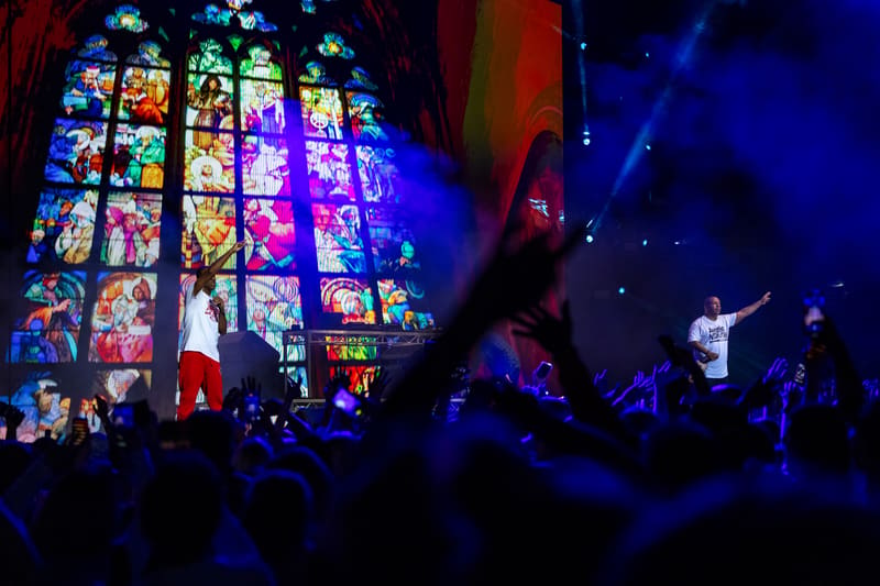 Large illuminated stained glass window backdrop with silhouetted crowd and performers under blue stage lighting at a live music event.