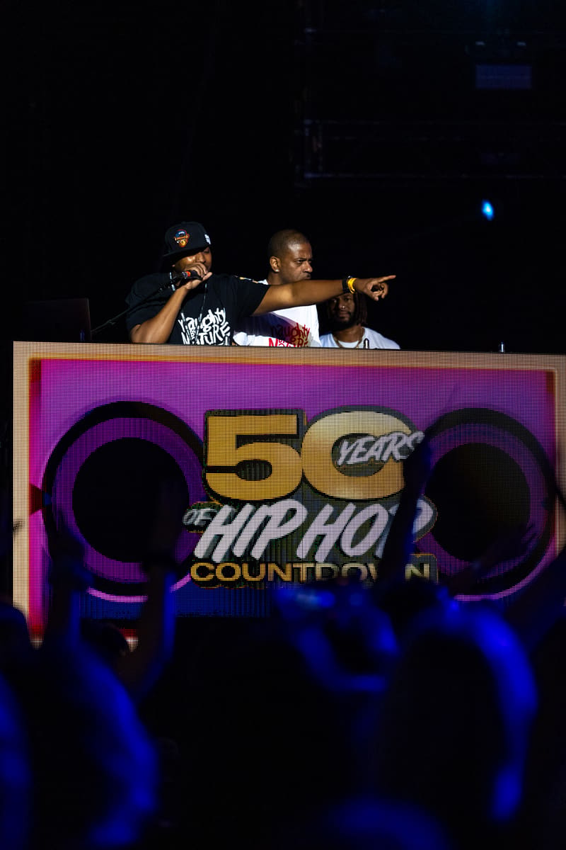 Two performers on stage at a hip-hop event, with one pointing to the crowd behind a colourful "50 Years of Hip Hop" banner.