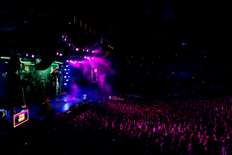 Large crowd watches performers on an illuminated stage bathed in purple and blue stage lighting.