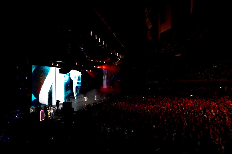 Large crowd faces an illuminated stage with performers silhouetted against blue and red lighting during a live event.