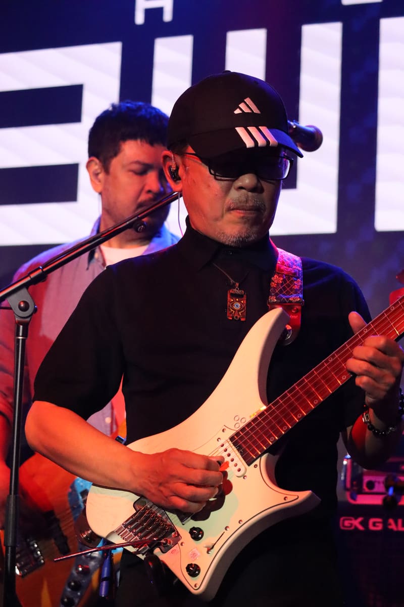 Guitarist in black cap and shirt playing white electric guitar under blue and purple stage lighting.