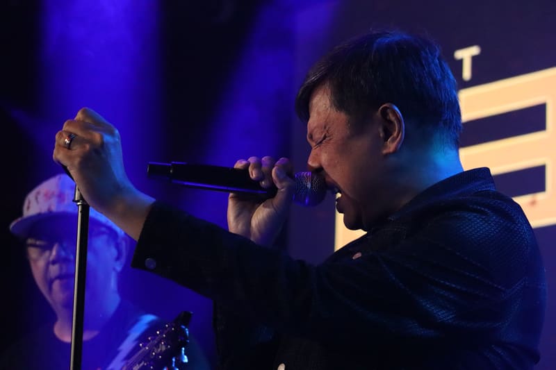 Two musicians perform on stage under blue lighting, one singing at a microphone stand while the other sings into a handheld microphone.