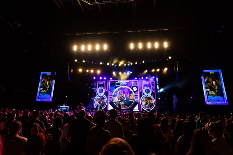Crowd of concert-goers with hands raised facing illuminated purple and gold stage setup with large screens and overhead lighting.