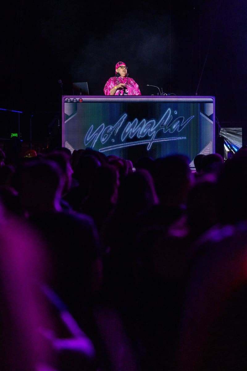 DJ in pink jacket performing at elevated booth with neon signage above silhouetted crowd bathed in purple stage lighting.