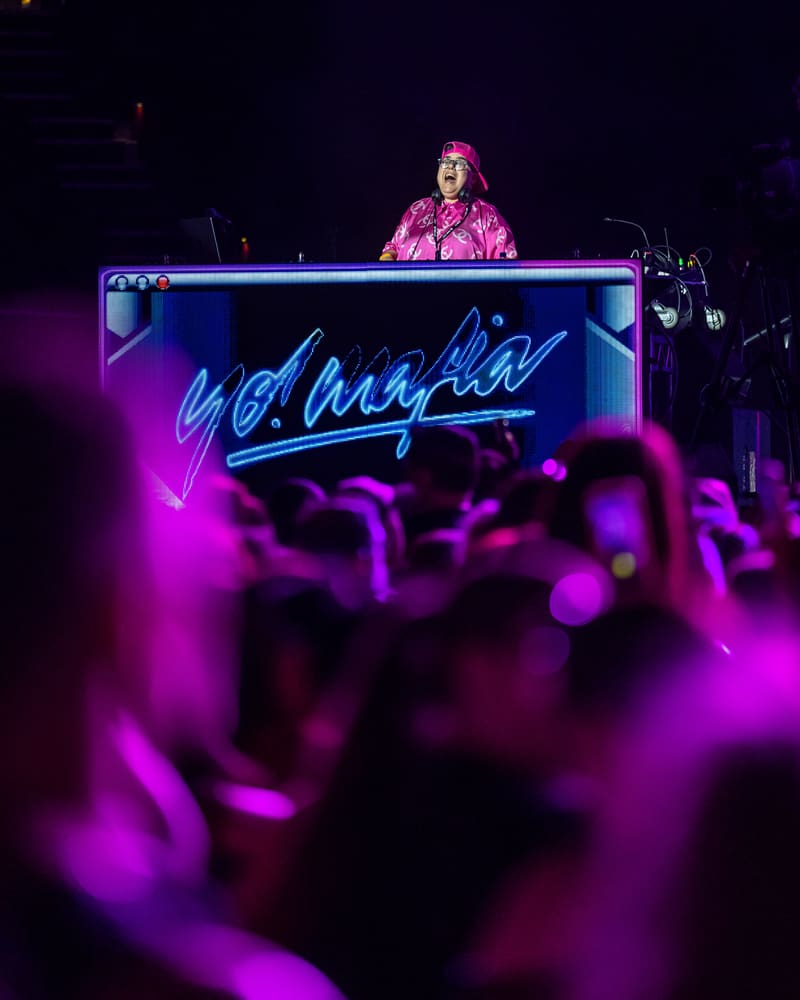 DJ performing at a neon-lit booth surrounded by a dancing crowd under purple stage lighting.