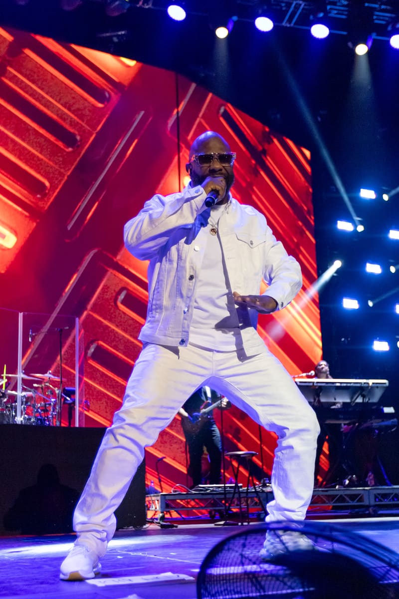 Male performer in white suit singing into microphone on stage with red neon backdrop and blue stage lighting.
