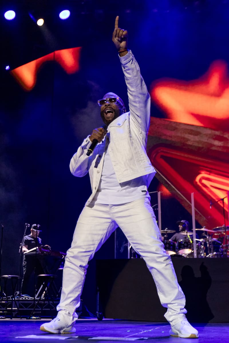 Male performer in white suit singing into microphone with fist raised, stage lit with blue and red lights.