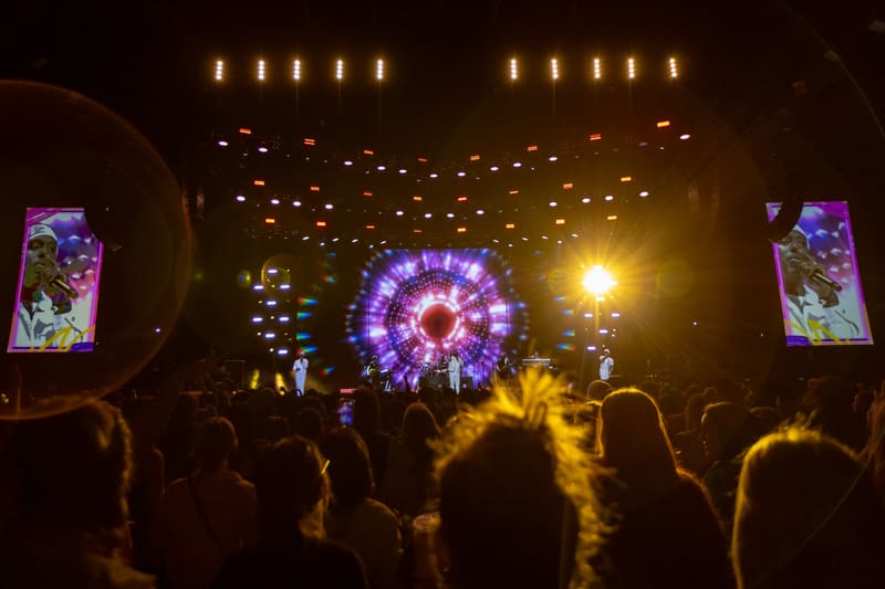 Audience members silhouetted against a vibrant purple and gold stage setup with large screens and dramatic lighting at a live concert venue.