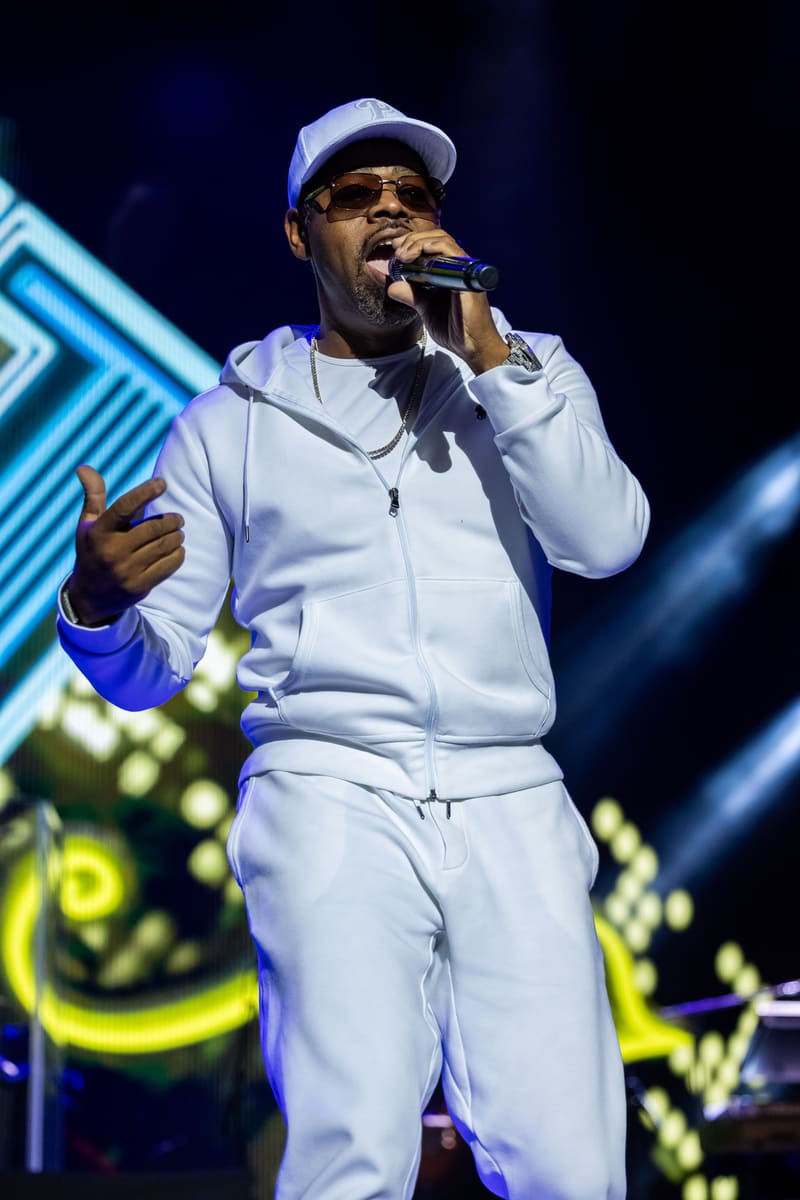 Performer in white tracksuit and cap singing into microphone under blue stage lighting during Fridayz Live concert.